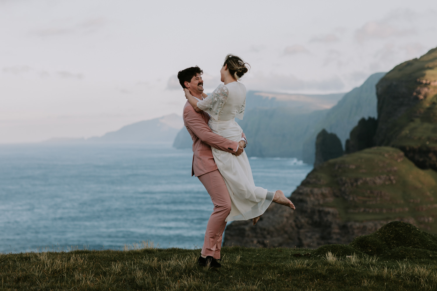 A man wearing a pink suit picks up his partner in a white wedding dress with dramatic sea cliffs in the background