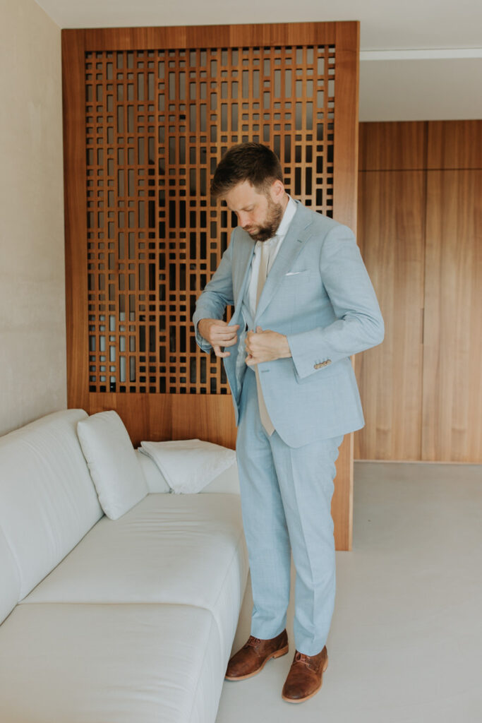 A man buttons his baby blue colored suit in a modern hotel room on his elopement wedding day