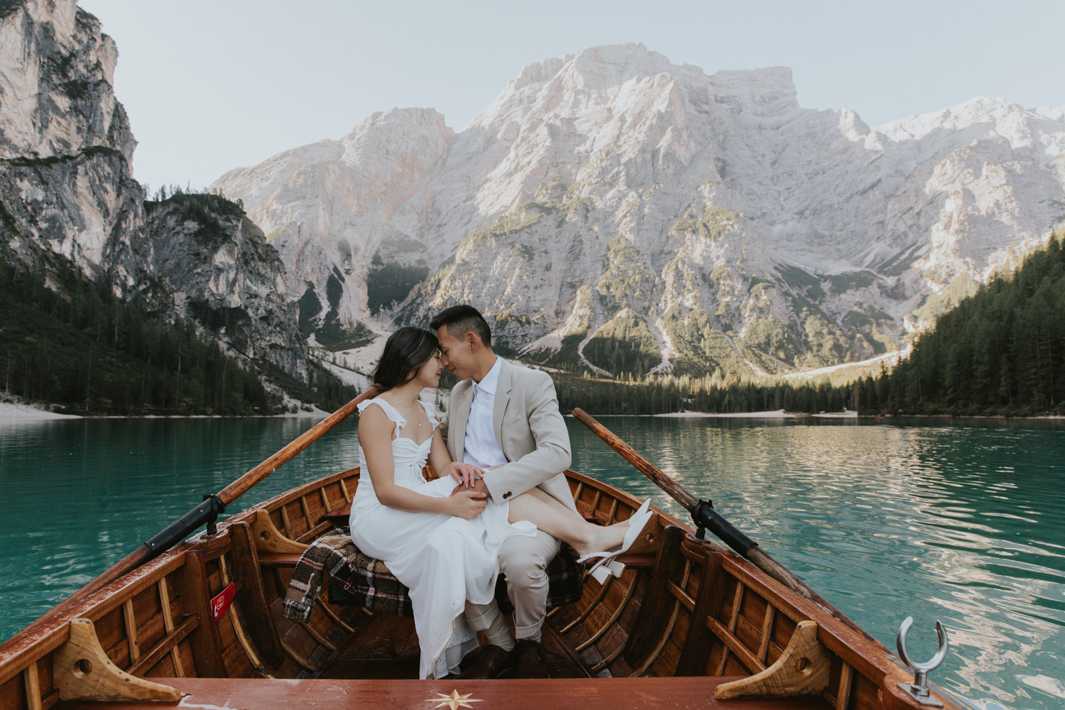 A couple sit face to face in a wooden boat on the famous Lago di Braies in the Italian Dolomites. The water around them is blue green and the mountains behind them are lit by sunlight.