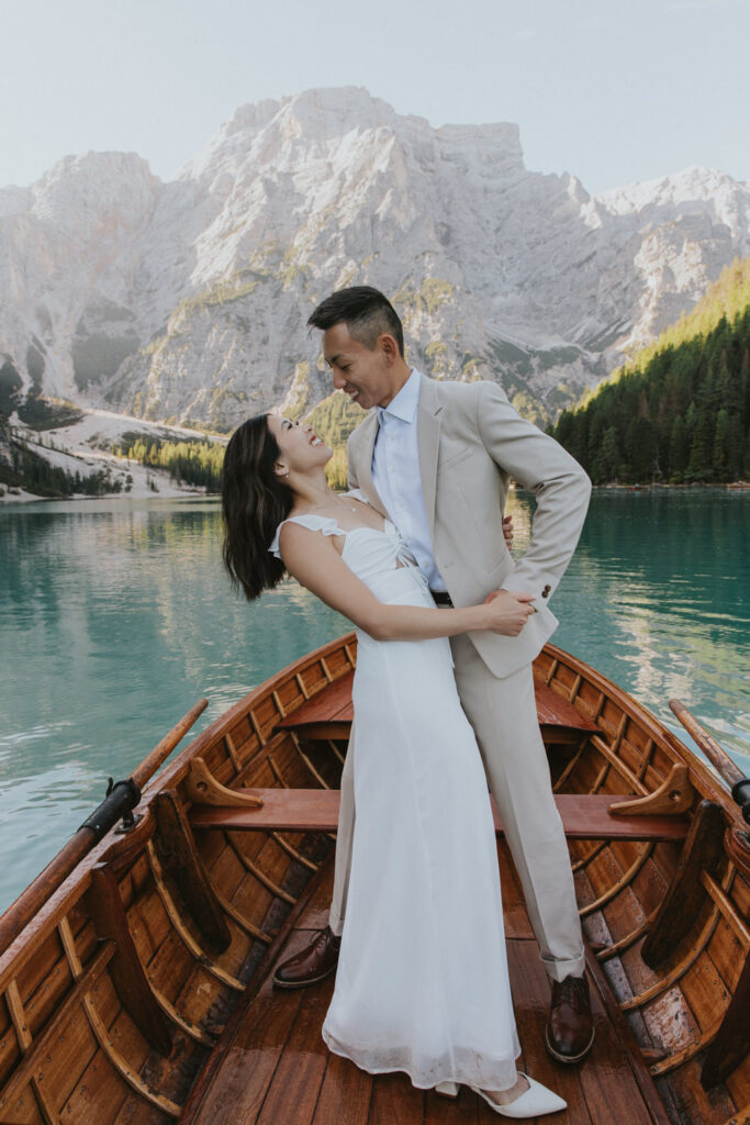 A wedding couple stand on a wooden boat at Lago di Braies during their Dolomites elopement wearing light creme colored suit and white dress