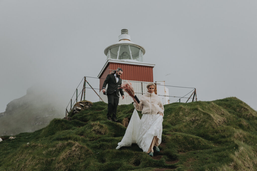 A couple in wedding attire walk down a narrow lighthouse trail with the dramatic cliffs of the Faroe Islands behind them