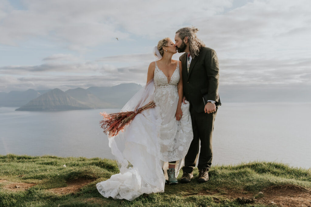 A couple in wedding attire stand on a sunny cliff kissing with the dramatic fjords of the Faroe Islands behind them