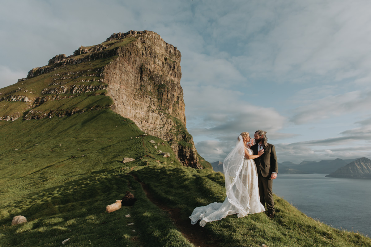 A couple in wedding attire stand on a sunny cliff with the dramatic fjords of the Faroe Islands behind them