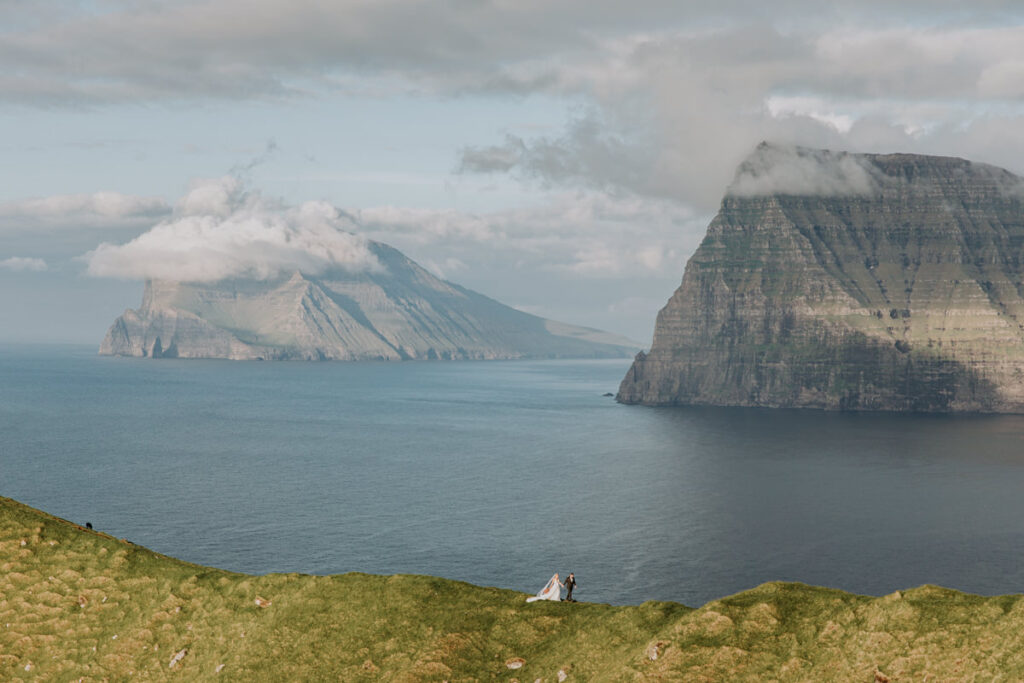 A couple in wedding attire walk far in the distance with the towering cliffs of the Faroe Islands across the fjords behind them
