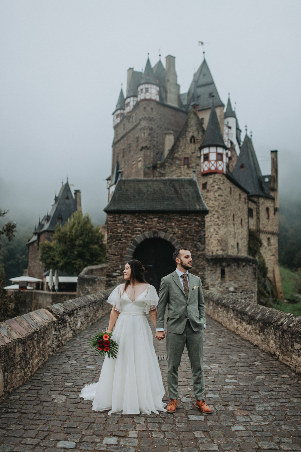 A couple stand in front of Burg Eltz during their elopement in Germany. The man is wearing an olive green suit with plaid tie and brown dress shoes.