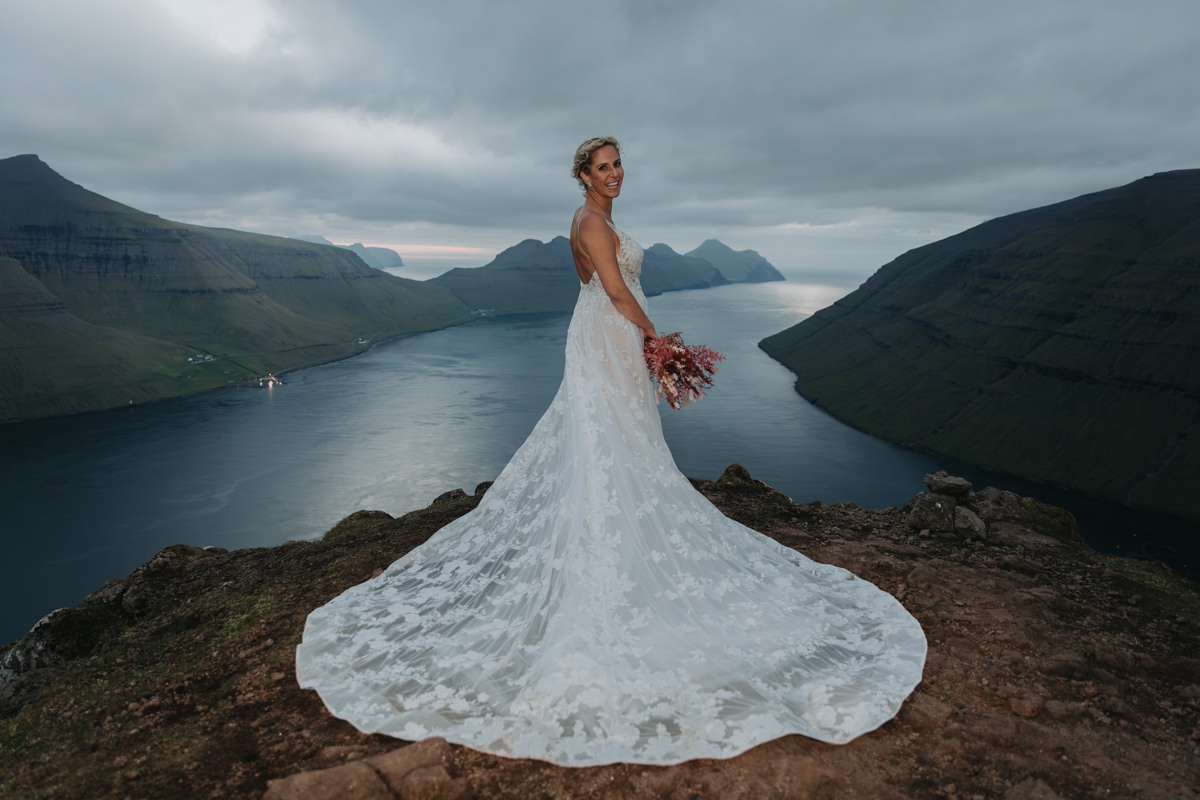 A woman in a sprawling wedding dress stands on a rocky cliff with the dramatic fjords of the Faroe Islands behind her