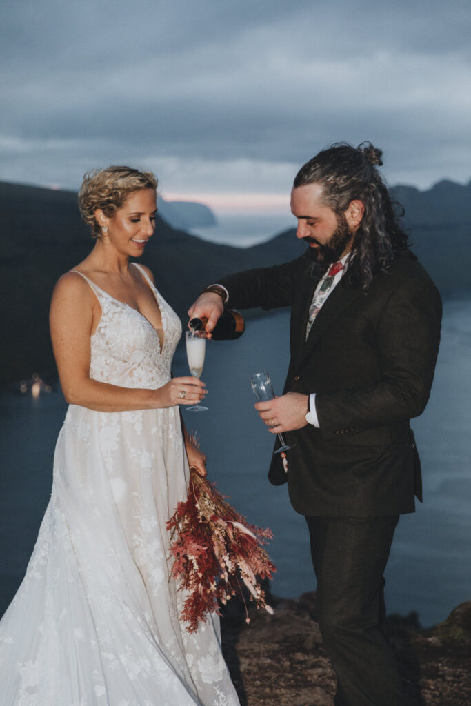 A couple in wedding attire pour champagne on a cliff with the dramatic fjords of the Faroe Islands behind them