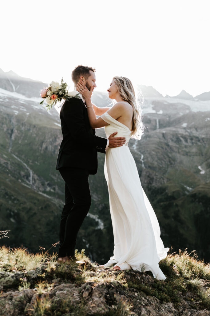 A couple in wedding clothes stand embracing with the sun behind them somewhere in a mountain valley in the Hohe Tauern area of Austria