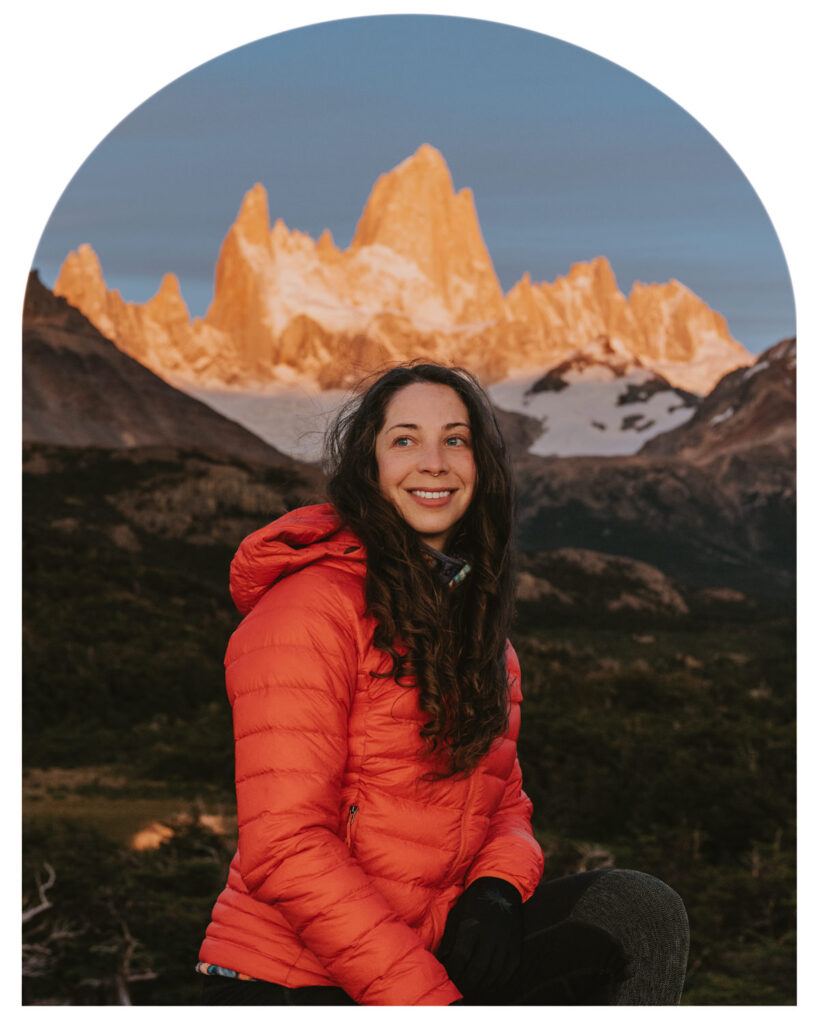 A white, female elopement photographer smiles in front of the camera wearing a bright red puffy. The iconic Fitz Roy mountain peak of Argentinean Patagonia is in the background.