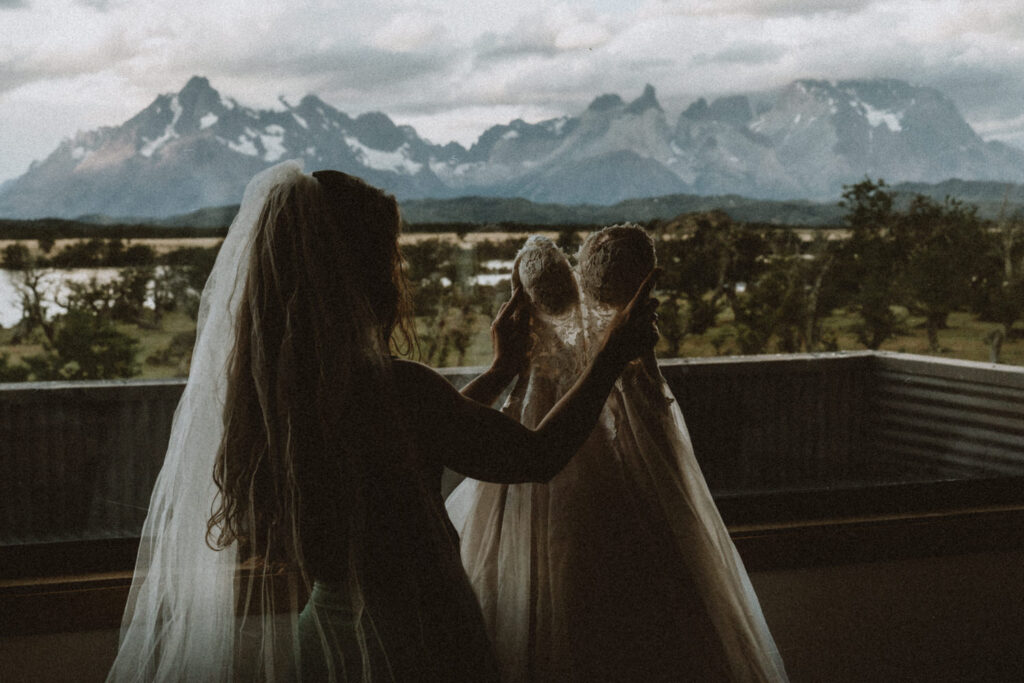 A woman in silhouette stands holding her wedding dress in front of a window during her Patagonia elopement. The Los Cuernos mountain is in the distance behind her. 