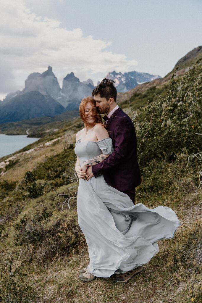 A couple in a dress and suit stand holding each other against the wind with jagged mountain peaks behind them. They are eloping in Patagonia.
