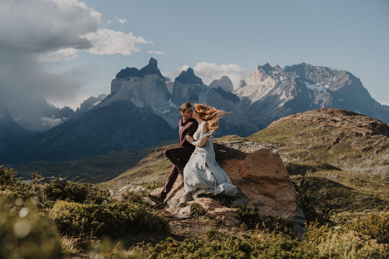 A couple sit in formal clothes posing for a Patagonia elopement embrace in strong winds with the Mirador los Cuernos mountain peaks behind them. They are eloping in Torres del Paine National Park in Chile on a sunny but very windy day.