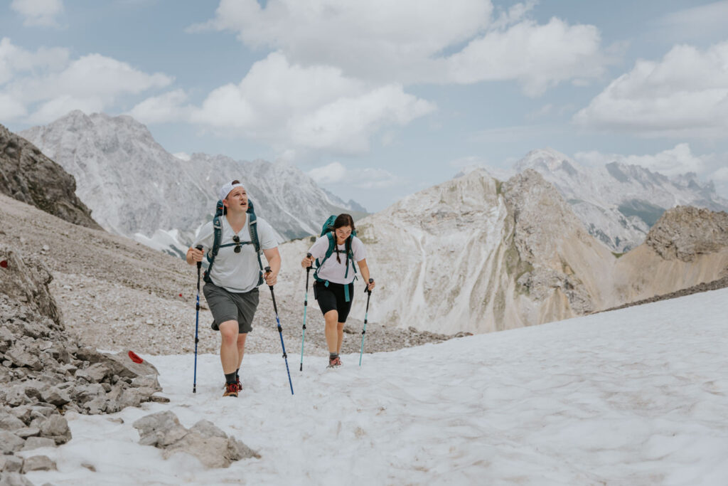 A couple hiking on their elopement day walk over snow on a pass near the Austrian Zugspitze, tall grey and green mountains behind them.