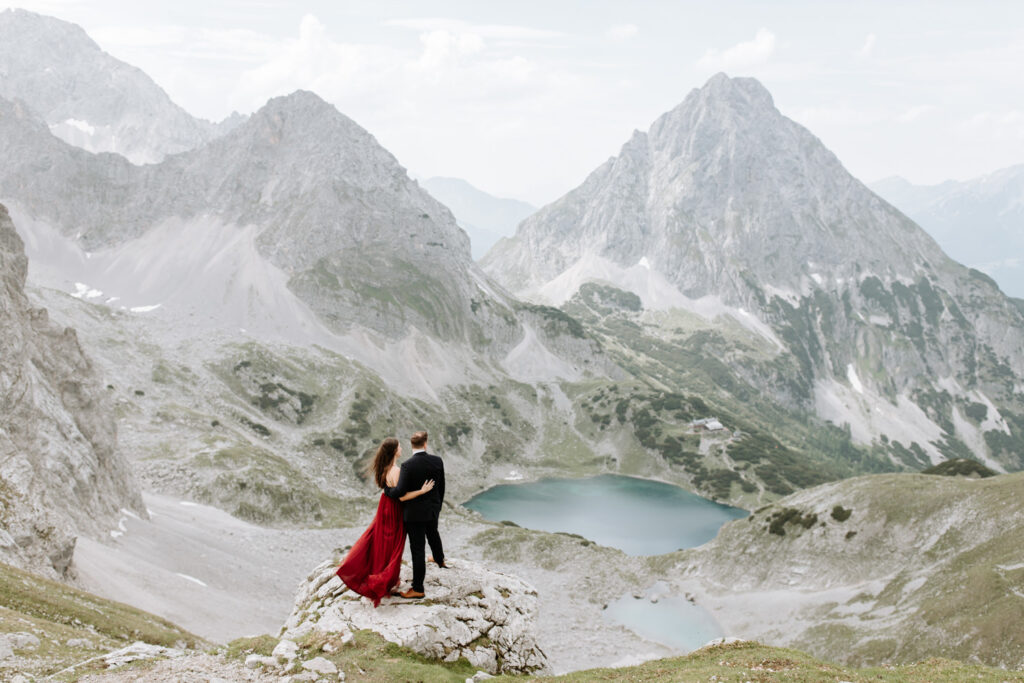 A couple hiking on their elopement day stand near the Austrian Zugspitze, a dramatic grey mountain scape in front of them and deep blue lakes below. The woman is wearing a red flowing dress and him a dark suit.