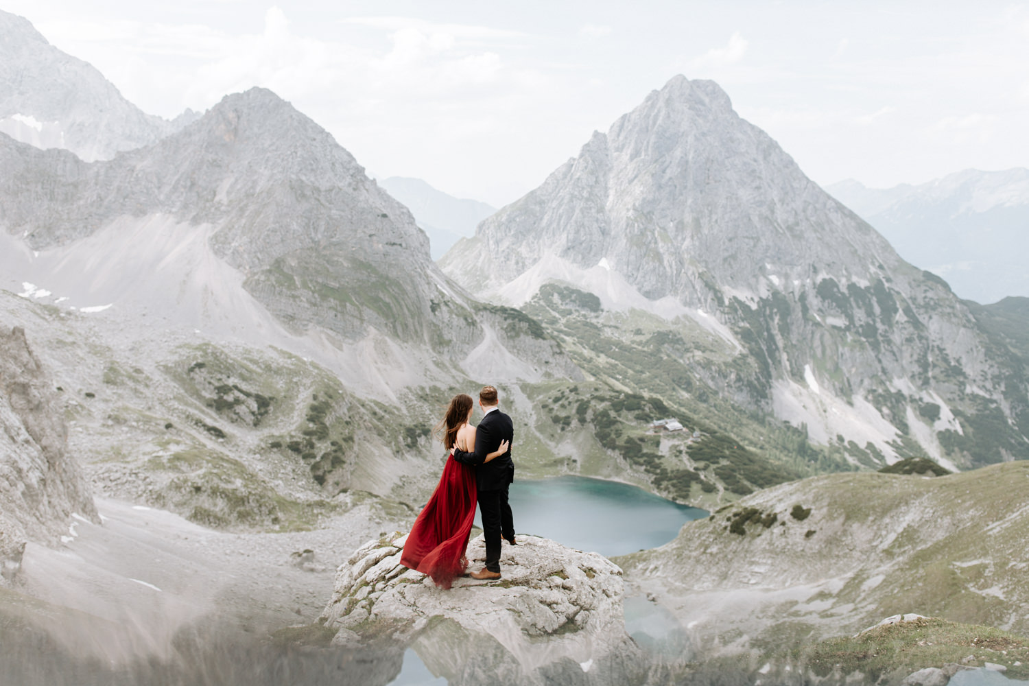 A couple hiking on their elopement day stand near the Austrian Zugspitze, a dramatic grey mountain scape in front of them and deep blue lakes. She is wearing a red flowing dress and him a dark suit.