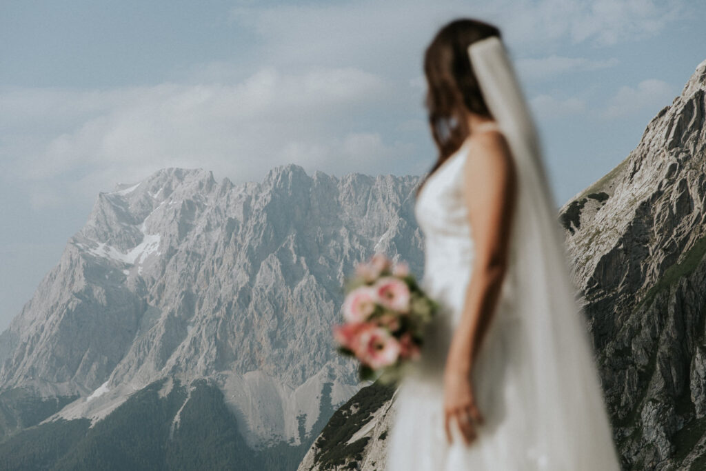 A bride hiking on her elopement day stands out of focus looking at the towering Austrian Zugspitze peak, holding pink wildflowers
