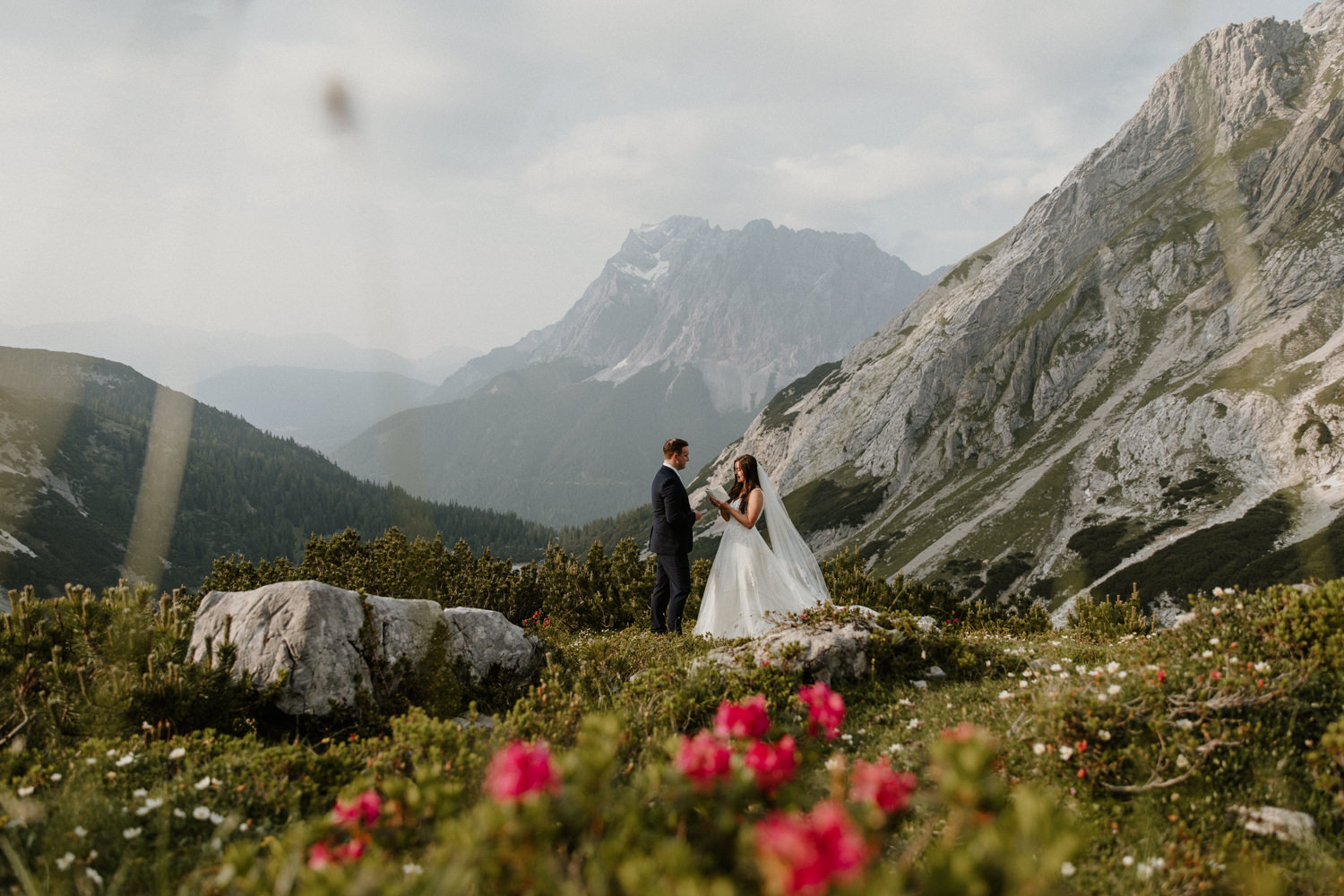 A couple hiking on their elopement day stand under the Austrian Zugspitze, framed by pink wildflowers, reading their wedding vows