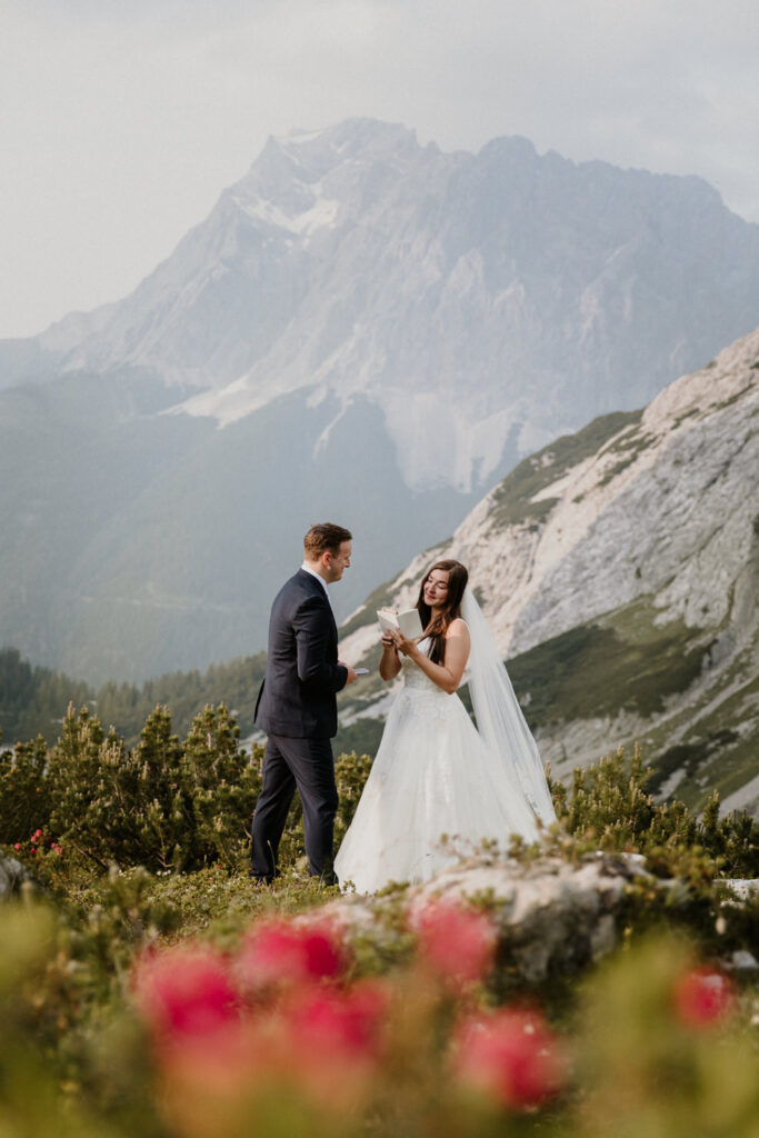 A couple read vows on their elopement day under the Austrian Zugspitze, framed by hot pink wildflowers.