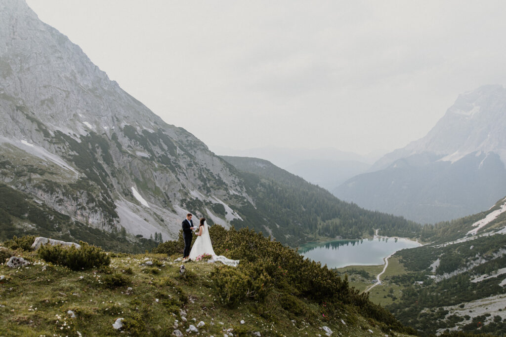 A couple hiking on their elopement day stand under the Austrian Zugspitze, framed by yellow and white wildflowers and a green lake, reading their vows.