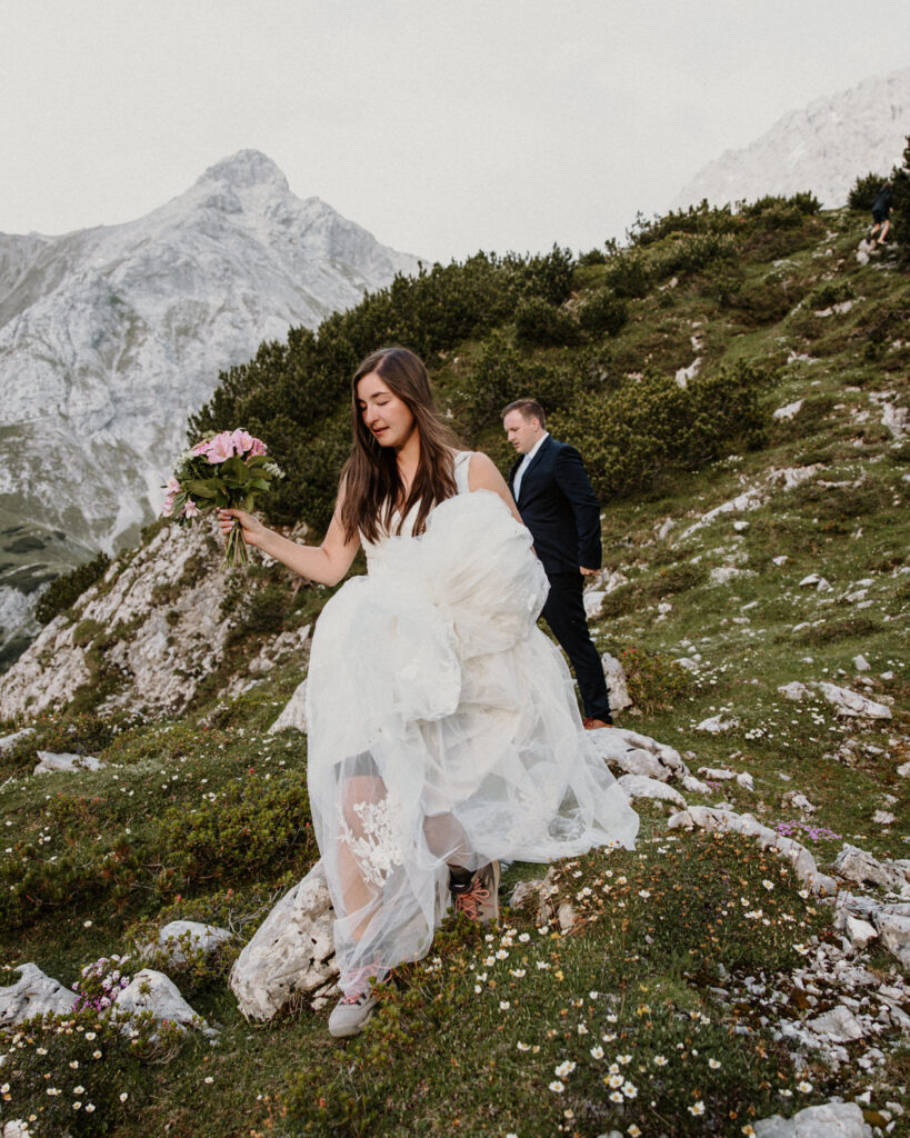 A couple hiking on their elopement day dance in wedding clothes under the Austrian Zugspitze, framed by white wildflowers and greenery.
