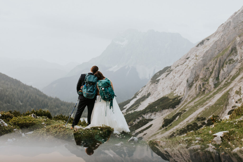 A couple hiking on their elopement day stand shoulder to shoulder facing the Austrian Zugspitze, wearing wedding clothes.
