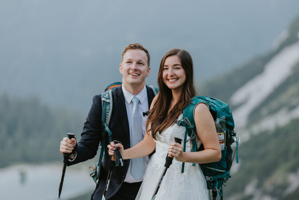 A couple hiking on their elopement day stand under the Austrian Zugspitze, posing for the camera with big smiles and holding hiking poles.