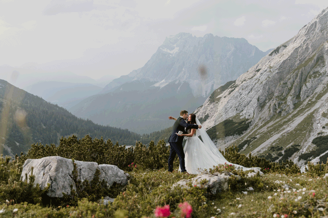 A couple hiking on their elopement day stand under the Austrian Zugspitze, framed by pink wildflowers and swaying grasses, reading their wedding vows.