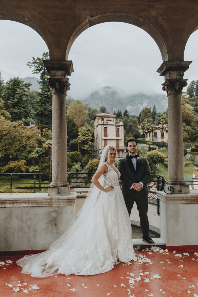 A wedding couple pose in white dress and green tux for their elopement photos on Lake Como with a villa and green trees in the background.