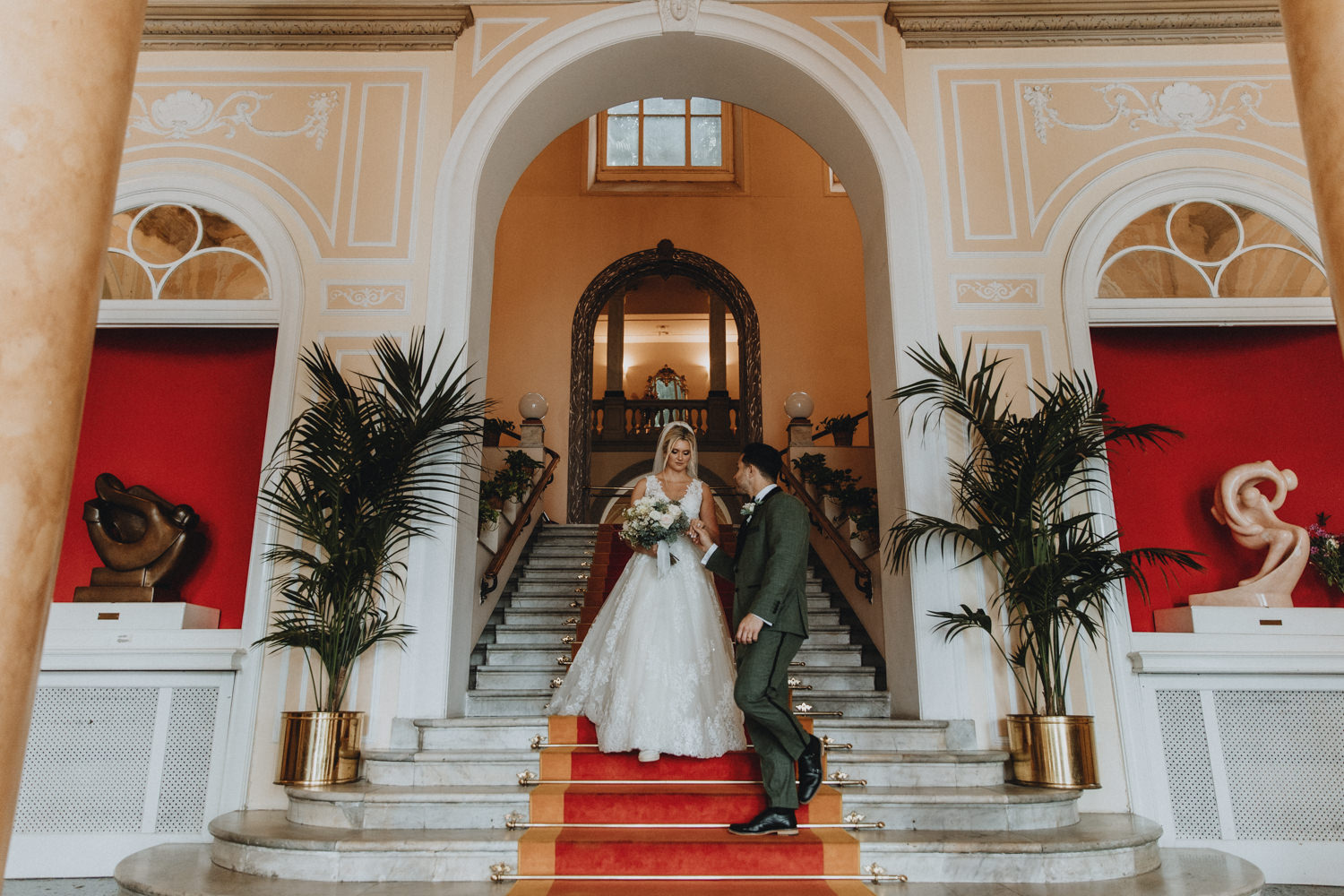 A couple in wedding outfits walk down the red and gold staircase in Grand Hotel Cadenabbia on their elopement day.