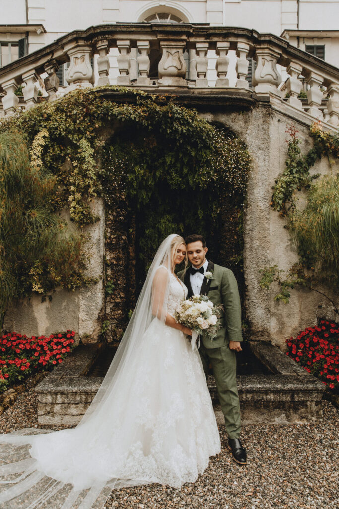 A couple pose at Villa Carlotta for their lake Como elopement. They are framed by red flowers, fountains, and draping greenery in the villa gardens.