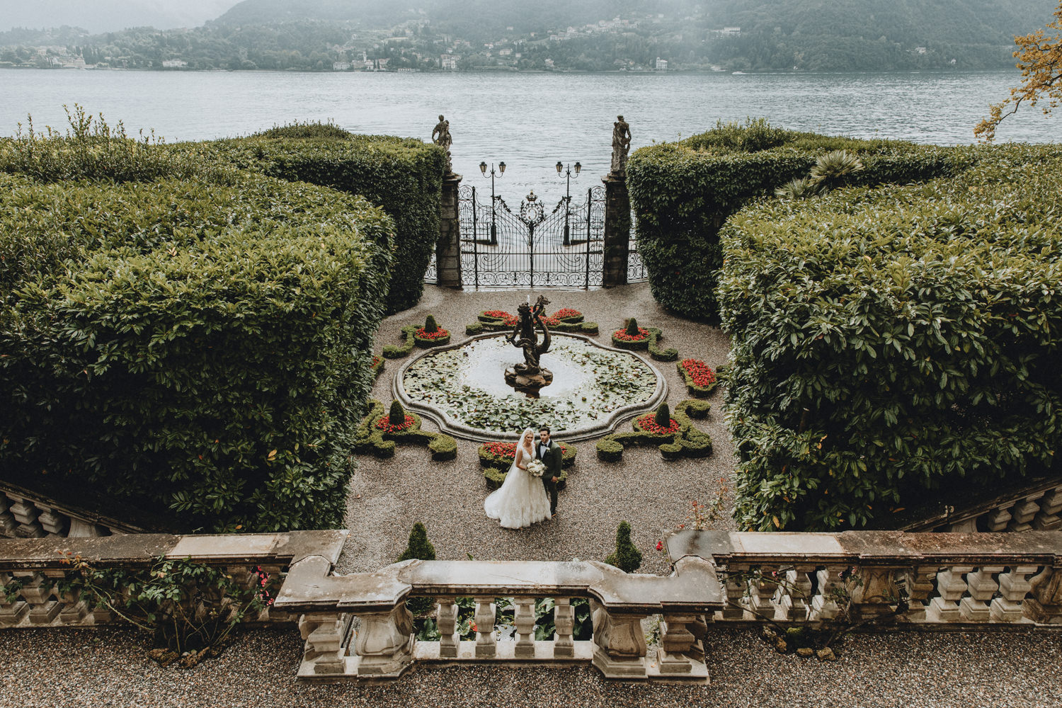 A couple pose far off in the frame at Villa Carlotta for their lake Como elopement. They are framed by red flowers, fountains, statues and bushes with the lake in the background.