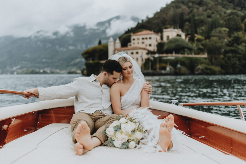 A couple dressed in wedding whites sits cuddling at the back of a speedboat at a lake in the European Alps