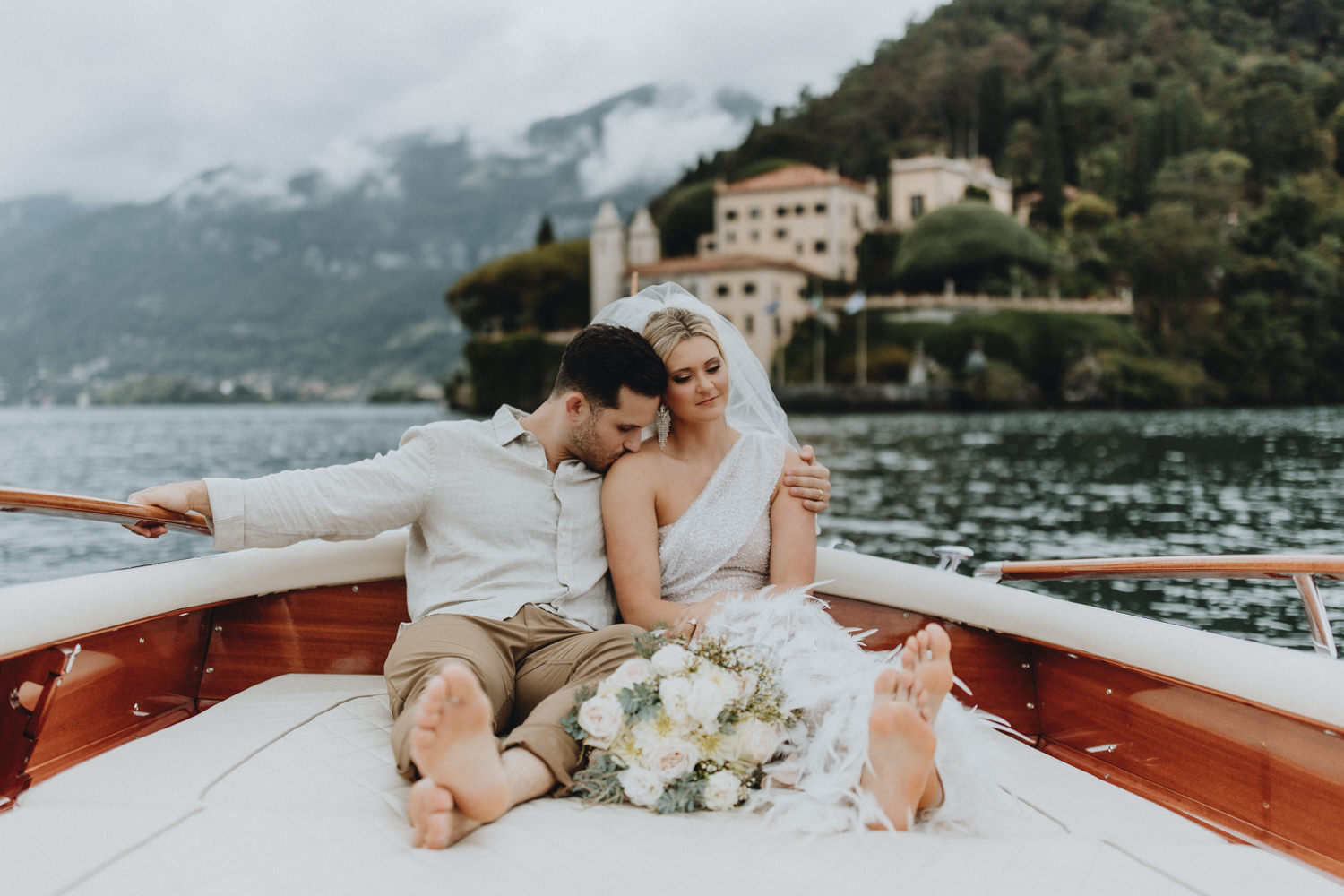 A couple in wedding attire sit on the back of a boat on Lake Como with a bouquet between them, cuddling in front of the impressive Villa del Balbianello