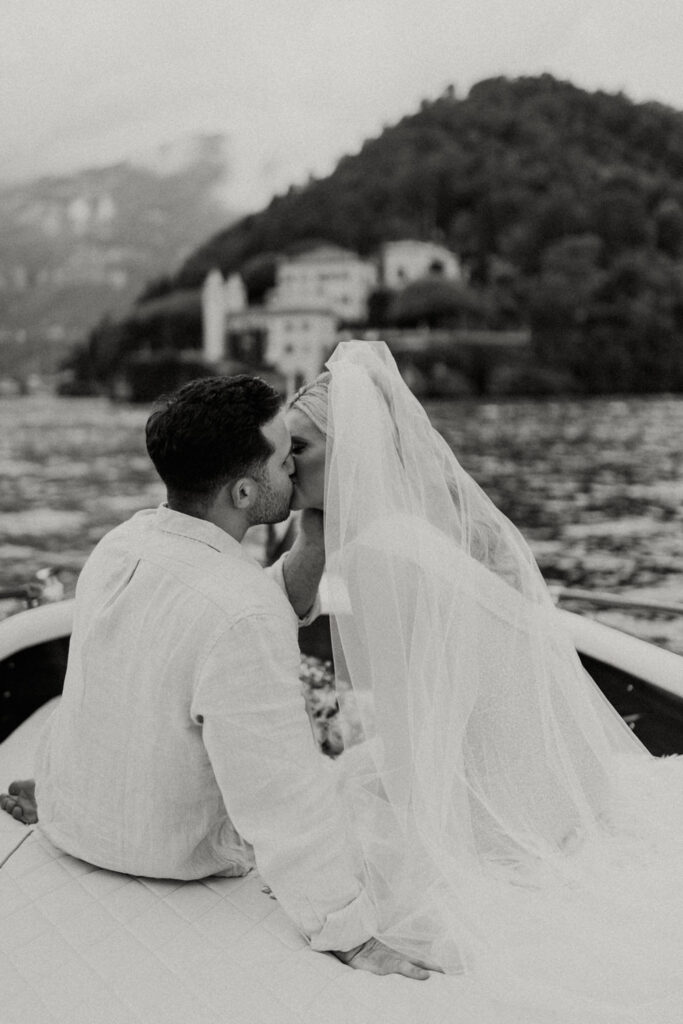 A couple in wedding attire sit on the back of a boat on Lake Como kissing and cuddling in front of the impressive Villa del Balbianello