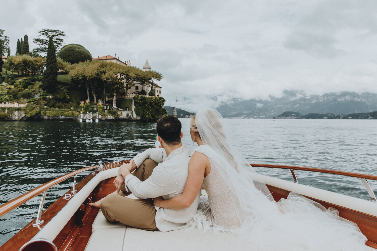 A couple in wedding attire sit on the back of a boat on Lake Como cuddling in front of the impressive Villa del Balbianello