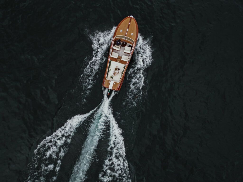 A classic wooden boat is photographed from above, skimming the waters of Lake Como with a bride and groom in the center near Villa del Balbianello