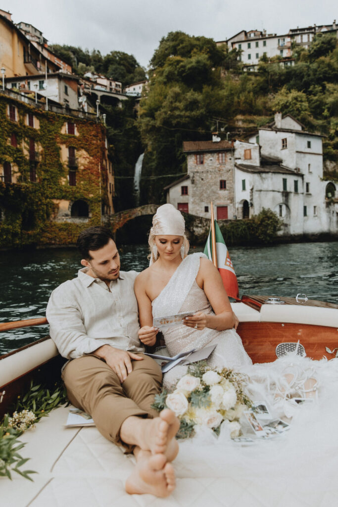 A couple in soft glam outfits sits on the back of a boat on Lake Como, framed by old Italian building, kissing as they read letters for their elopement day
