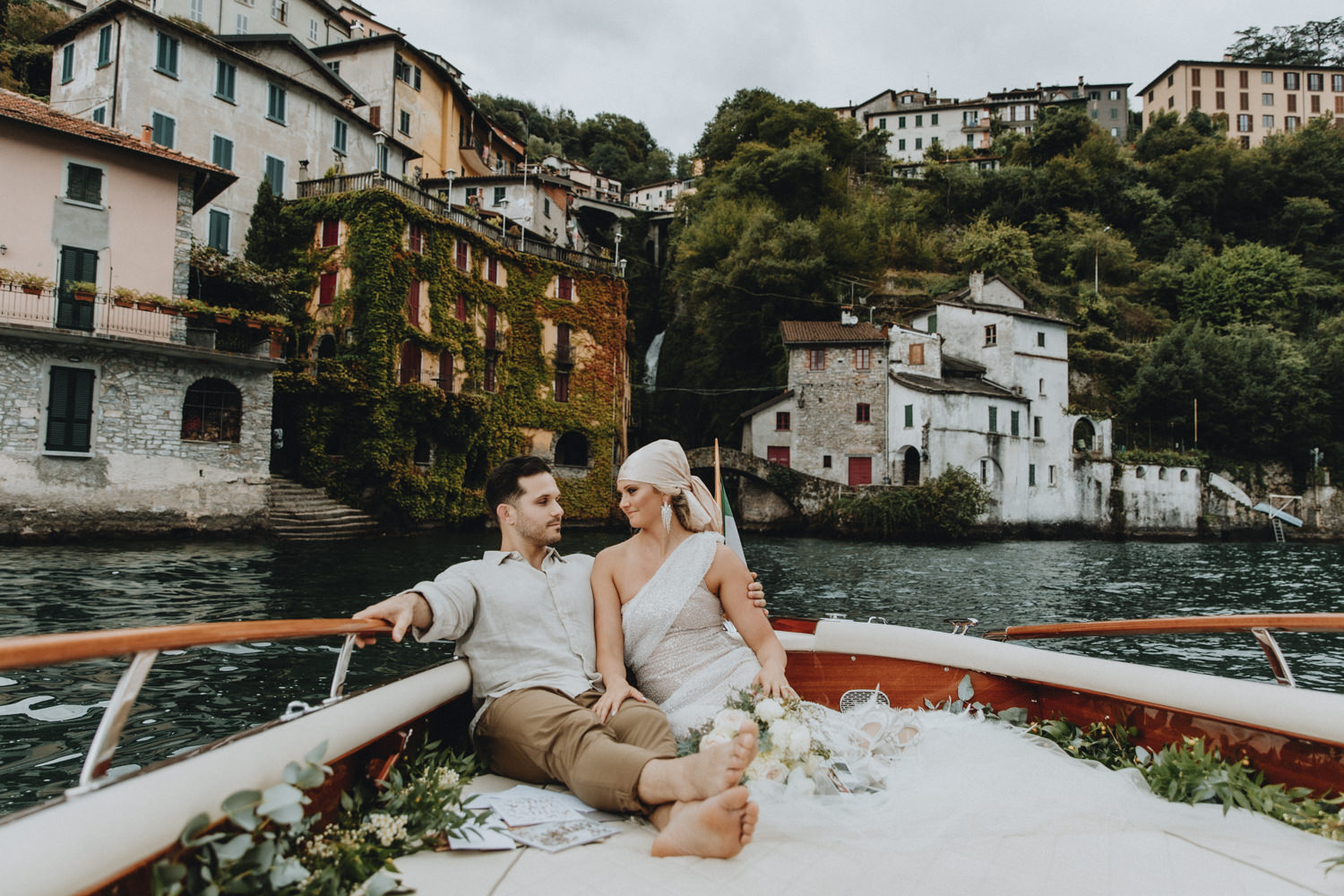 A couple in light toned glam oufits relax on the back of a classic wooden boat on Lake Como near the Nesso waterfall on their moody September elopement day
