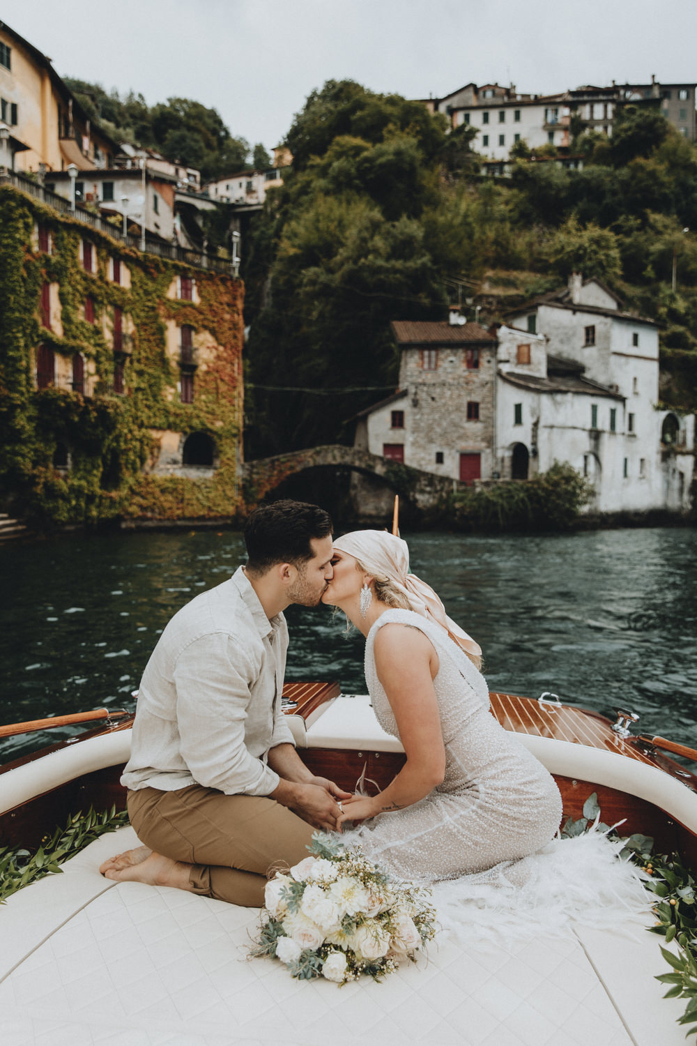 A couple kisses on the back of a wooden boat on lake Como during their elopement day abroad. The water is a deep blue and there are old Italian buildings and a waterfall on the land behind them.