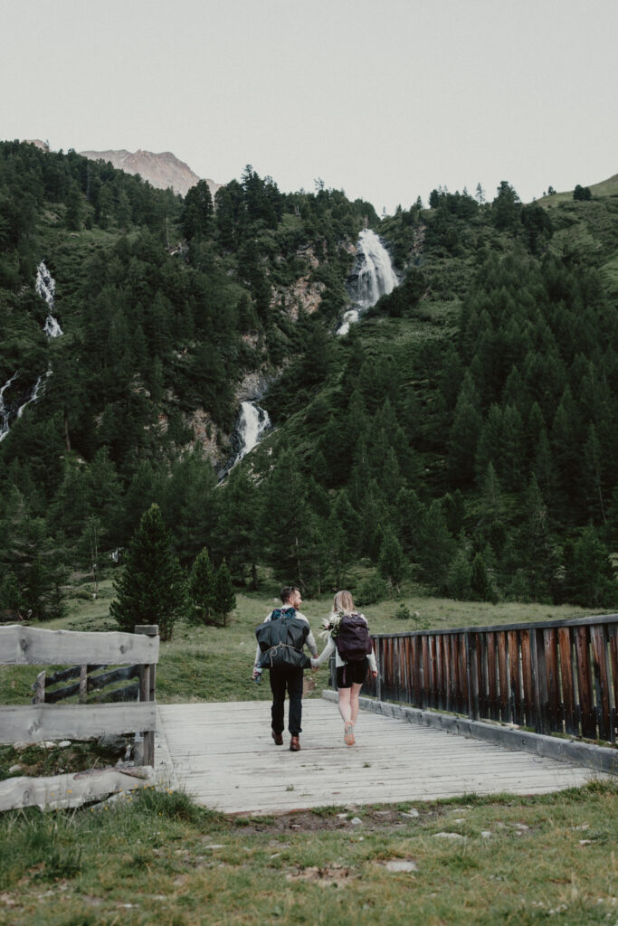 A couple are hiking in a mountain valley in Austria, walking across a wooden bridge toward a waterfall on an August morning. 