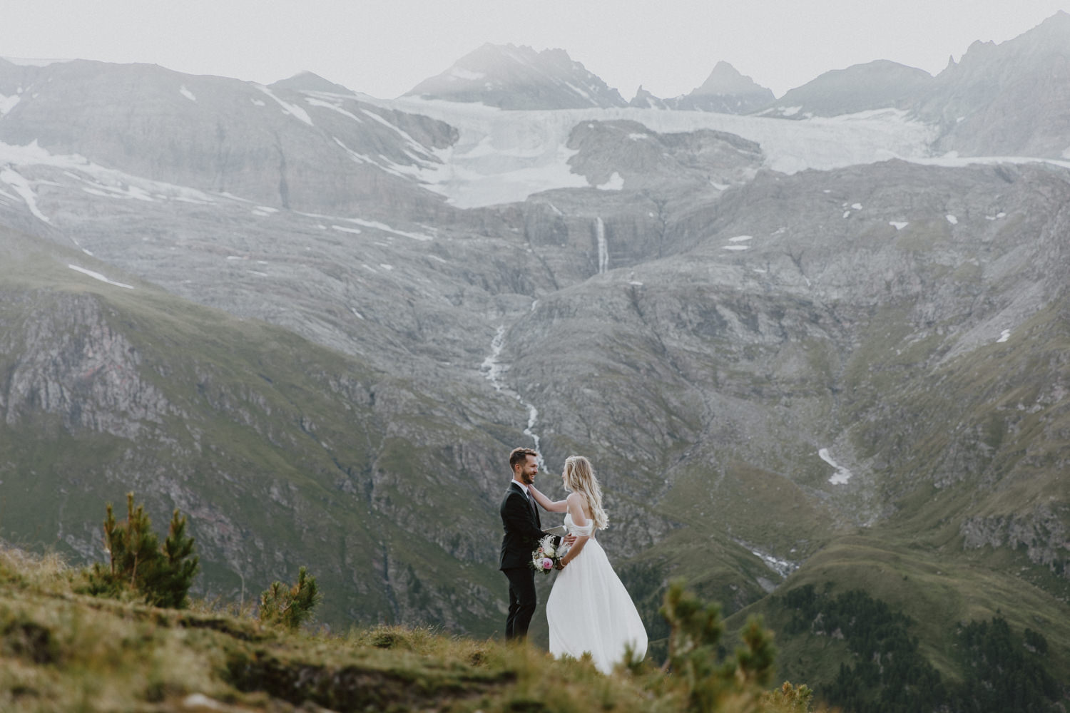 A couple are hiking on a green mountain with glaciers and waterfalls behind them in August. They are wearing wedding clothes and reading elopement vows.
