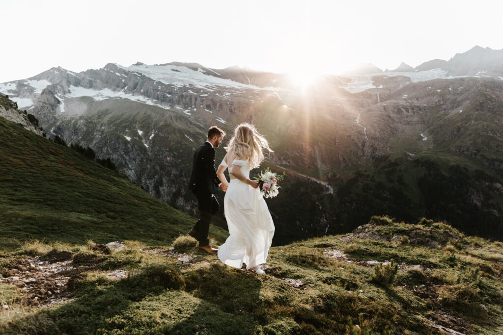A couple hiking in a white dress and black suit run away from the camera on a mountain toward a bright sun in August on their elopement day