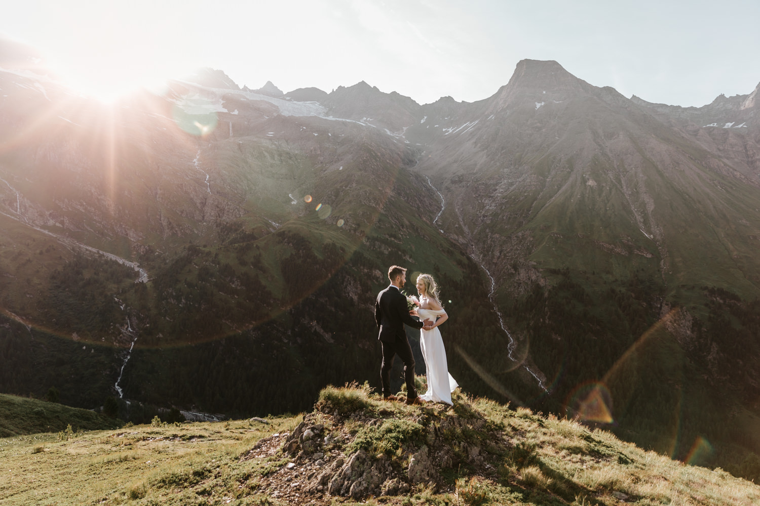 A couple hiking in a white dress and black suit stand on a mountain in August with a bright sun flaring above the peaks on their elopement day