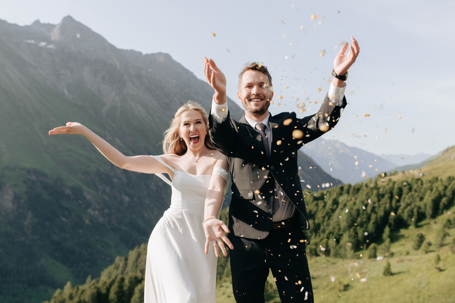 A white couple in white wedding dress and black suit laugh and toss native flowers on a mountaintop in Austria during their hiking elopement.
