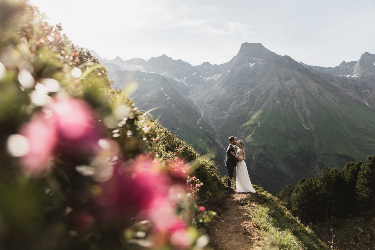 A couple stand small in the center of the frame with dramatic Austrian mountains in the background while hiking. They are framed by pink flowers in the foreground, celebrating their elopement.
