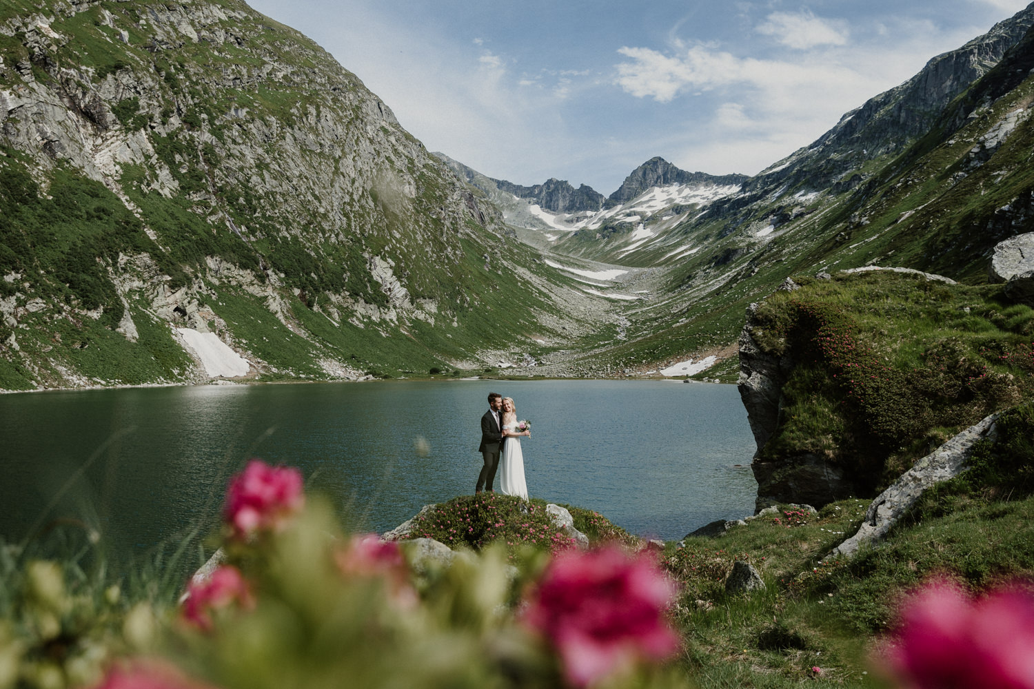 A couple stand small in the center of the frame with dramatic Austrian mountains and a blue lake in the background while hiking. They are framed by pink flowers in the foreground, celebrating their elopement.