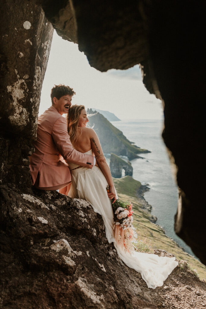 A hetero couple in white wedding dress and pink suit sit against a small cave opening, smiling with the dramatics islets of the Faroe Islands behind them.
