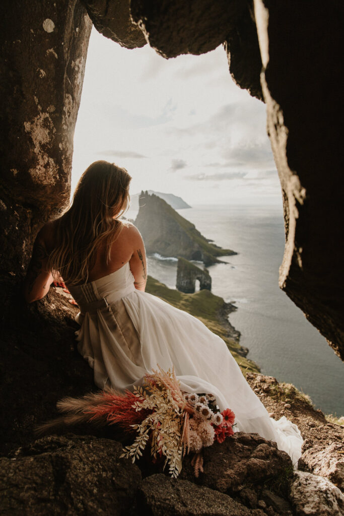 A bride in white wedding dress sits in front of a pink and white bouquet facing away from the camera toward a dramatic, golden sunset with the jagged peaks of the Faroe Islands in the distance
