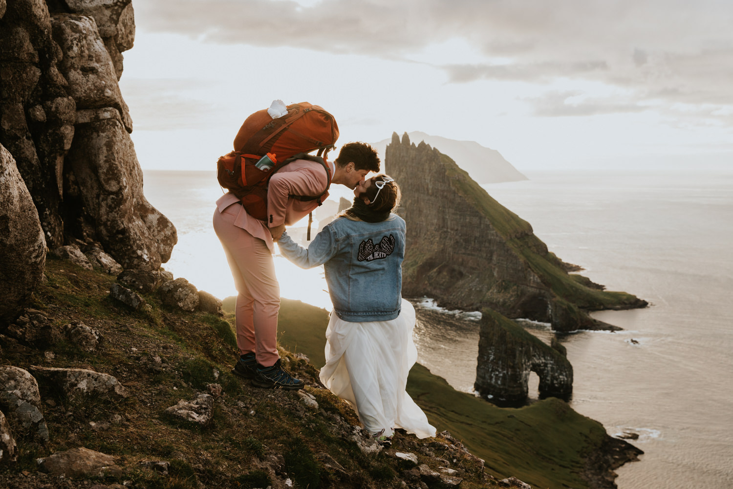 A hetero couple in white wedding dress and jean jacket, pink suit and backpack, kiss in front of the dramatic landscape of the Faroe Islands behind them.