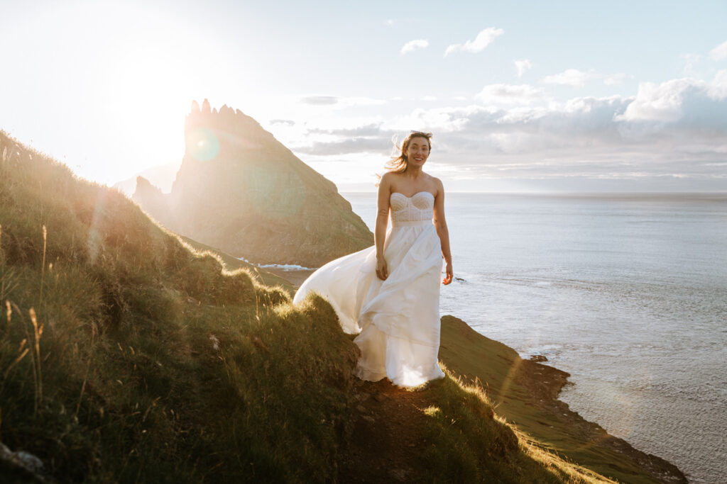 A blonde woman with white wedding dress smiles in front of a dramatic, evening sun and the jagged cliffs of the Faroe Islands behind her.